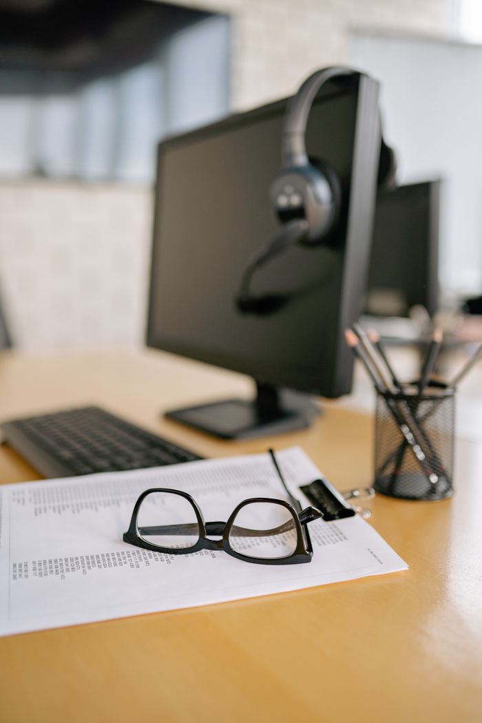 about-us-02 Eyeglasses on a paper next to a computer and headset on a wooden desk.